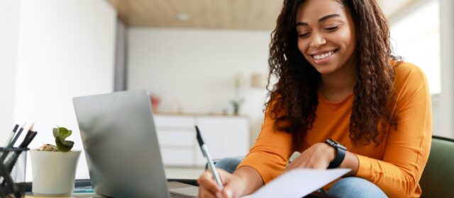 Black woman sitting at desk, using computer writing in notebook