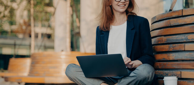 Pleased pretty redhead woman learns educational course on laptop computer sits crossed legs on wooden bench outdoors drinks takeaway coffee looks happily into distance creats new publication